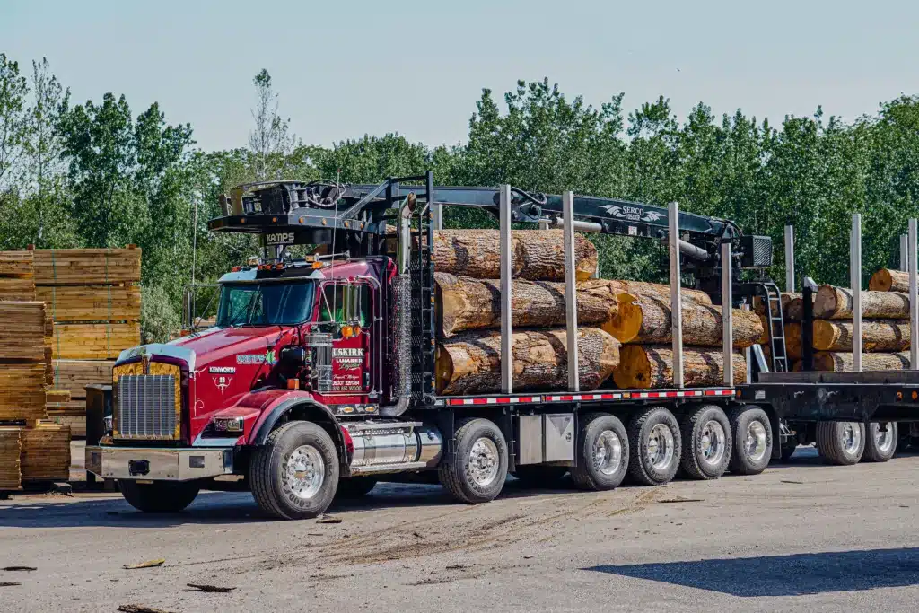 Red semi truck reading "Buskirk Lumber Logistics" is parked in the foreground. On its open trailer are large cut timber logs.